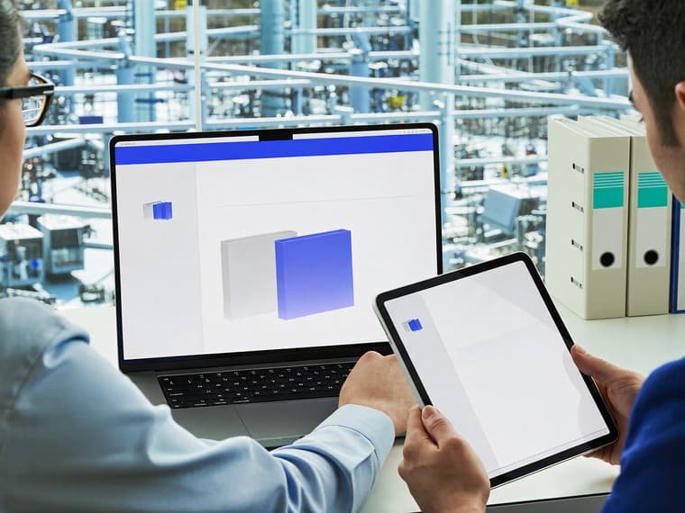 Two colleagues at a mezzanine desk comparing a document across a laptop and a tablet, a shelf of ring binders beside them.
