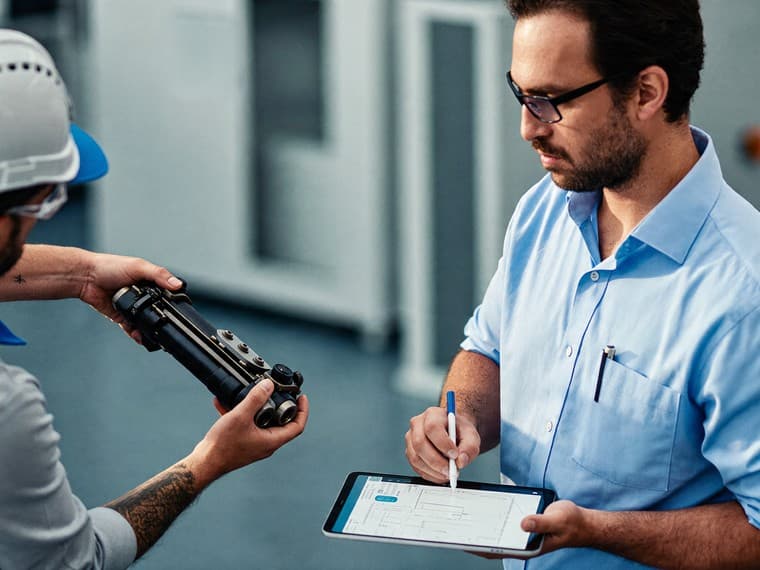 A supervisor writing on a tablet while a machine operator in a hard hat holds up the failed part he is describing.