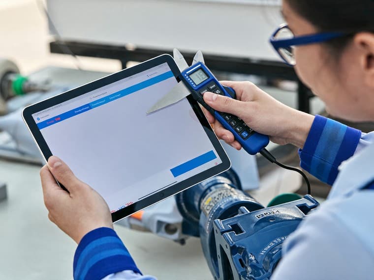 An inspector in safety glasses measuring a machined part with digital calipers, holding a tablet in her other hand.